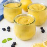 3 jars of lemon curd surrounded by lemon slices and blueberries.
