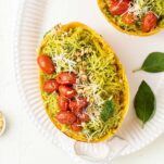 A spaghetti squash shown prepared with pesto, tomatoes, walnuts, and parmesan, on a white platter.