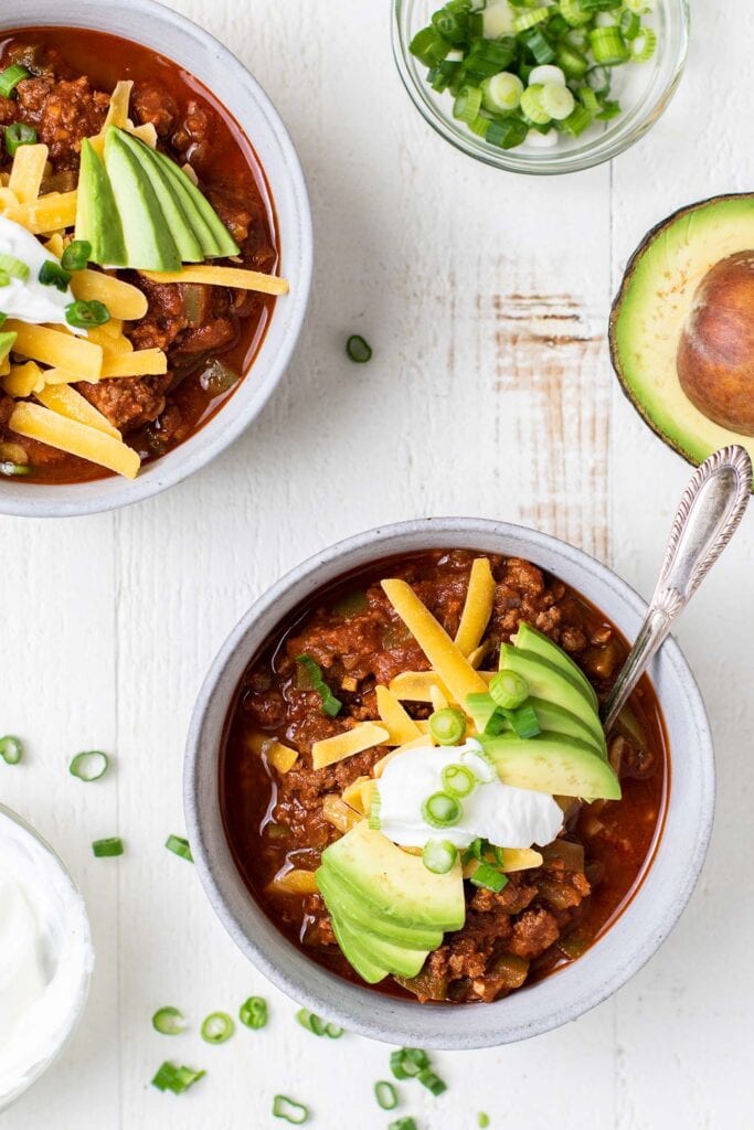 Two bowls of beef chili topped with cheese, avocado, sour cream, and green onions.