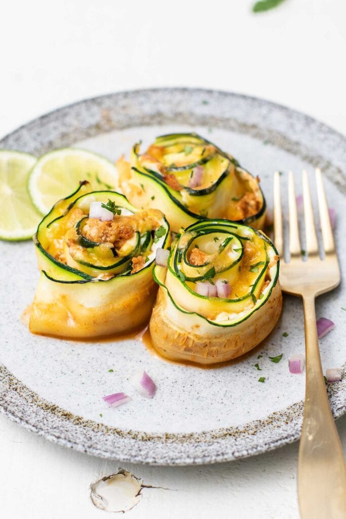 3 zucchini enchilada roll ups on a plate garnished with lime slices and cilantro.