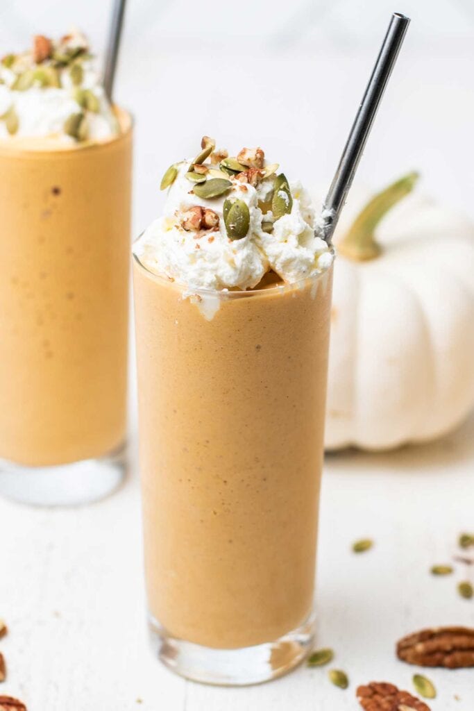 A pumpkin smoothie served in a tall glass garnished with whipped cream and pumpkin seeds.
