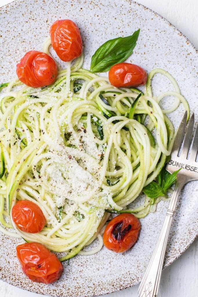 Zoodles shown cooked on a plate garnished with parmesan cheese, basil, and burst tomatoes.