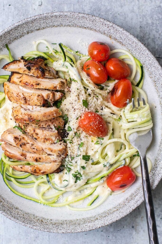 A plate with zoodles tossed with alfredo sauce, topped with chicken and burst tomatoes.