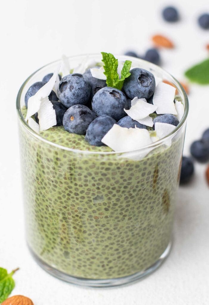 A glass filled with a vibrant green match chia pudding, topped with blueberries and coconut.