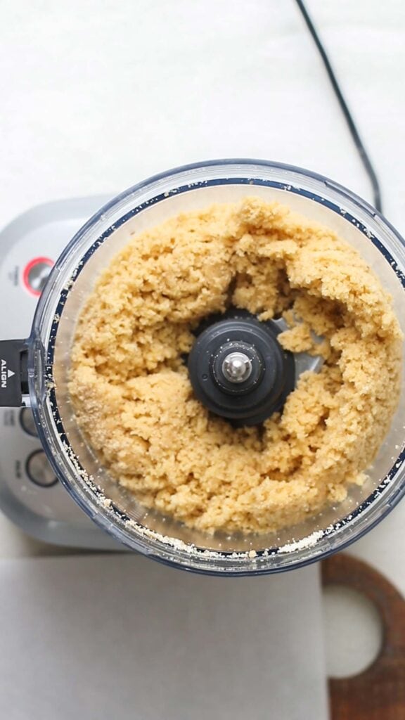 A food processor showing a crumbly dough in the bowl.